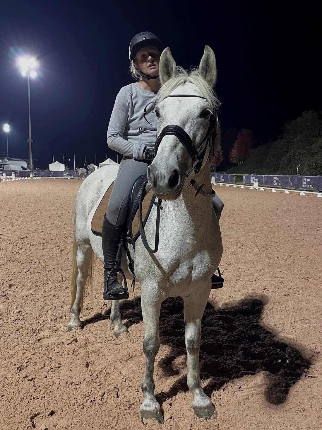Un cheval gris monté dans la carrière éclairée de l’écurie, à la nuit tombée.